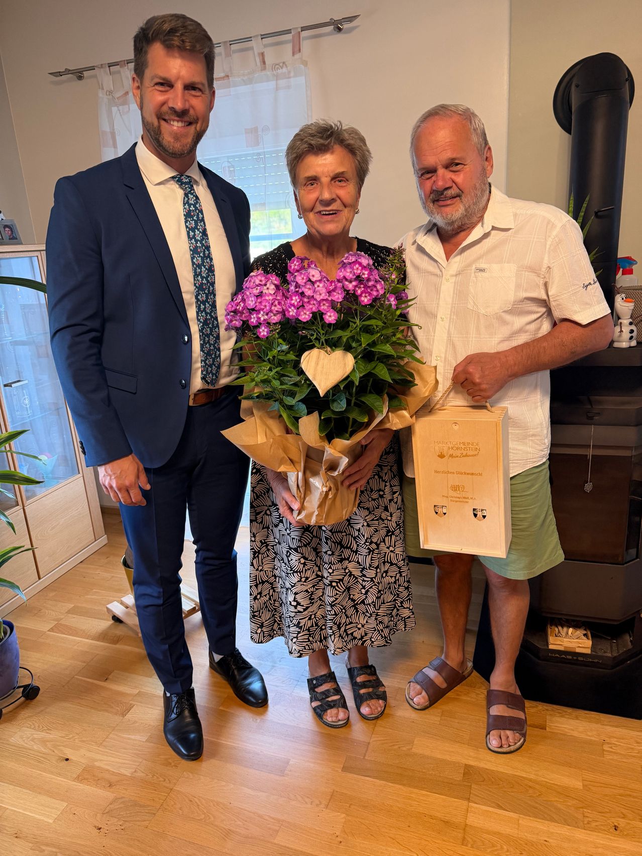 Drei Menschen stehen lächelnd in einem Raum. Die Frau in der Mitte hält einen Strauß lila Blumen und eine Geschenktüte. Der Mann links trägt einen Anzug und eine Krawatte, während der Mann rechts ein weißes Hemd und grüne Shorts trägt.