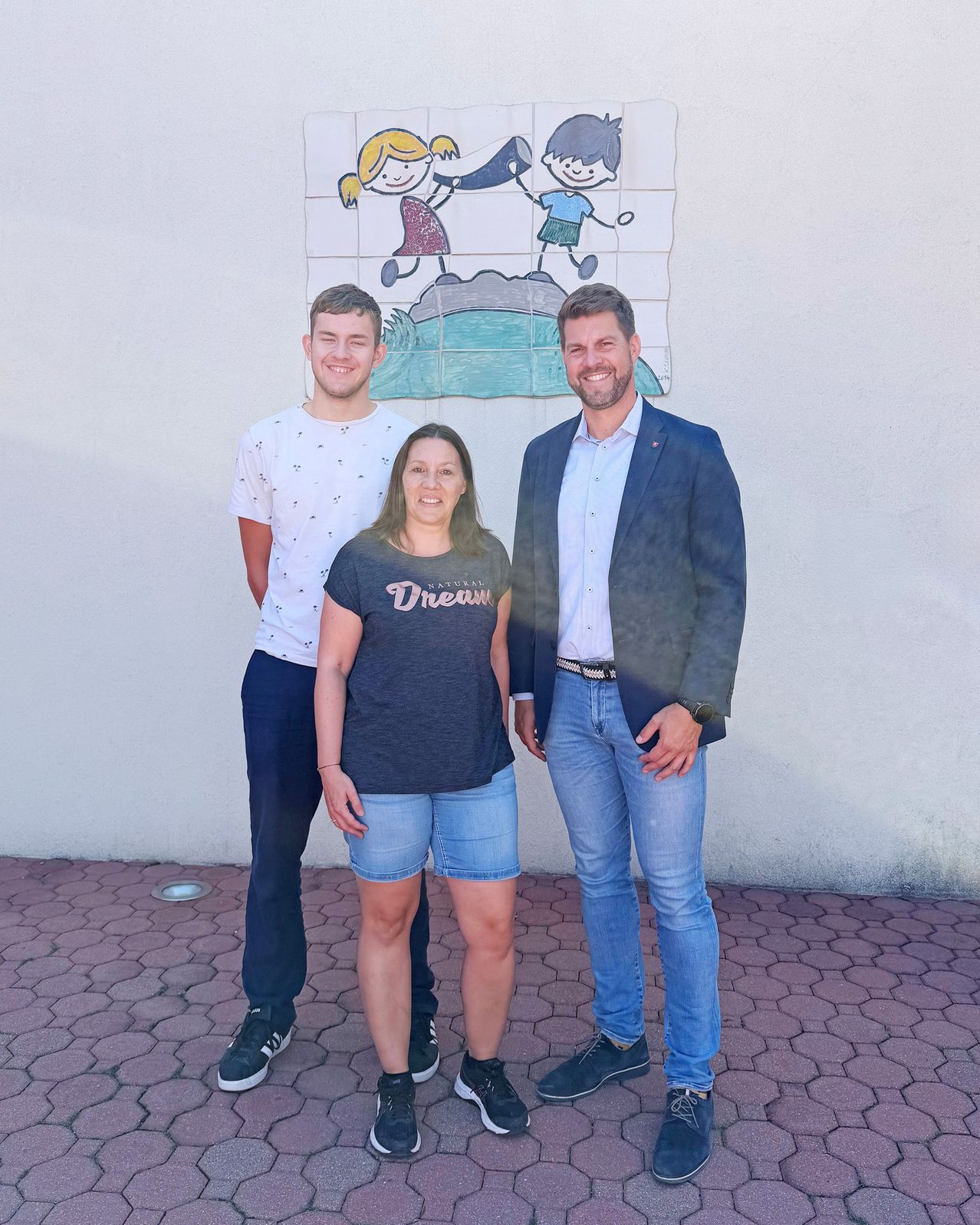 Drei Personen stehen vor einer Wand mit einem Wandbild von spielenden Kindern. Der Mann rechts trägt einen Anzug und eine Uhr. Die Frau in der Mitte trägt ein Shirt mit dem Wort 'Traum'. Der Mann links trägt ein weißes Shirt und Sneakers.