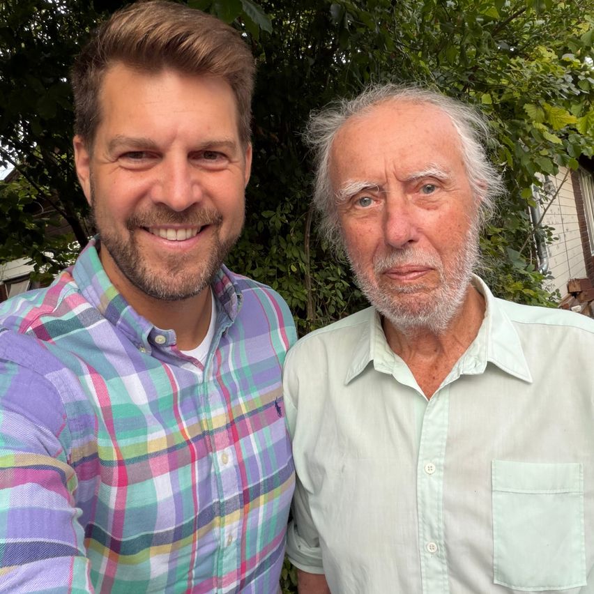 Zwei Männer stehen nebeneinander und lächeln für ein Foto. Der Mann auf der linken Seite trägt ein farbenfrohes kariertes Hemd, und der Mann auf der rechten Seite trägt ein hellgrünes Poloshirt. Sie stehen vor einem Baum mit grünen Blättern.