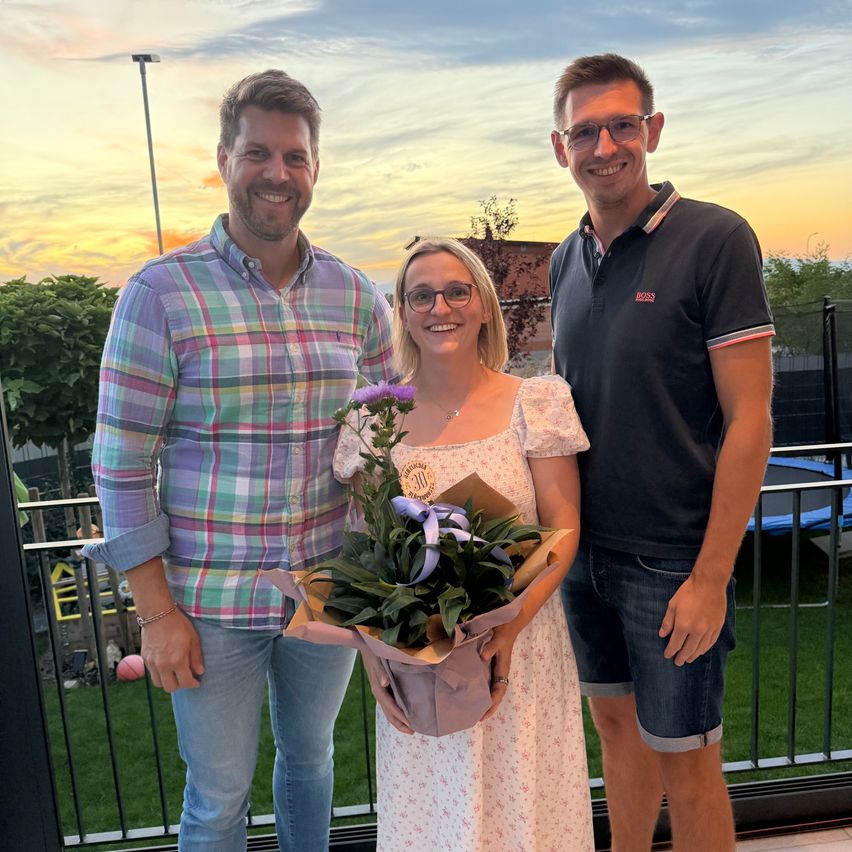 Drei Personen stehen bei Sonnenuntergang auf einem Balkon und lächeln. Eine Frau hält einen Blumenstrauß.
