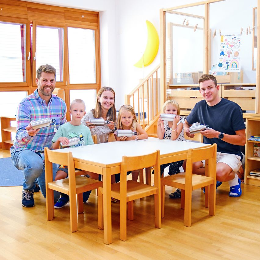 Eine Familie mit fünf Personen, darunter zwei Erwachsene und drei Kinder, sitzt um einen Tisch herum, lächelt und hält silberne zylindrische Objekte in den Händen. Der Raum ist hell mit Holzböden und großen Fenstern.