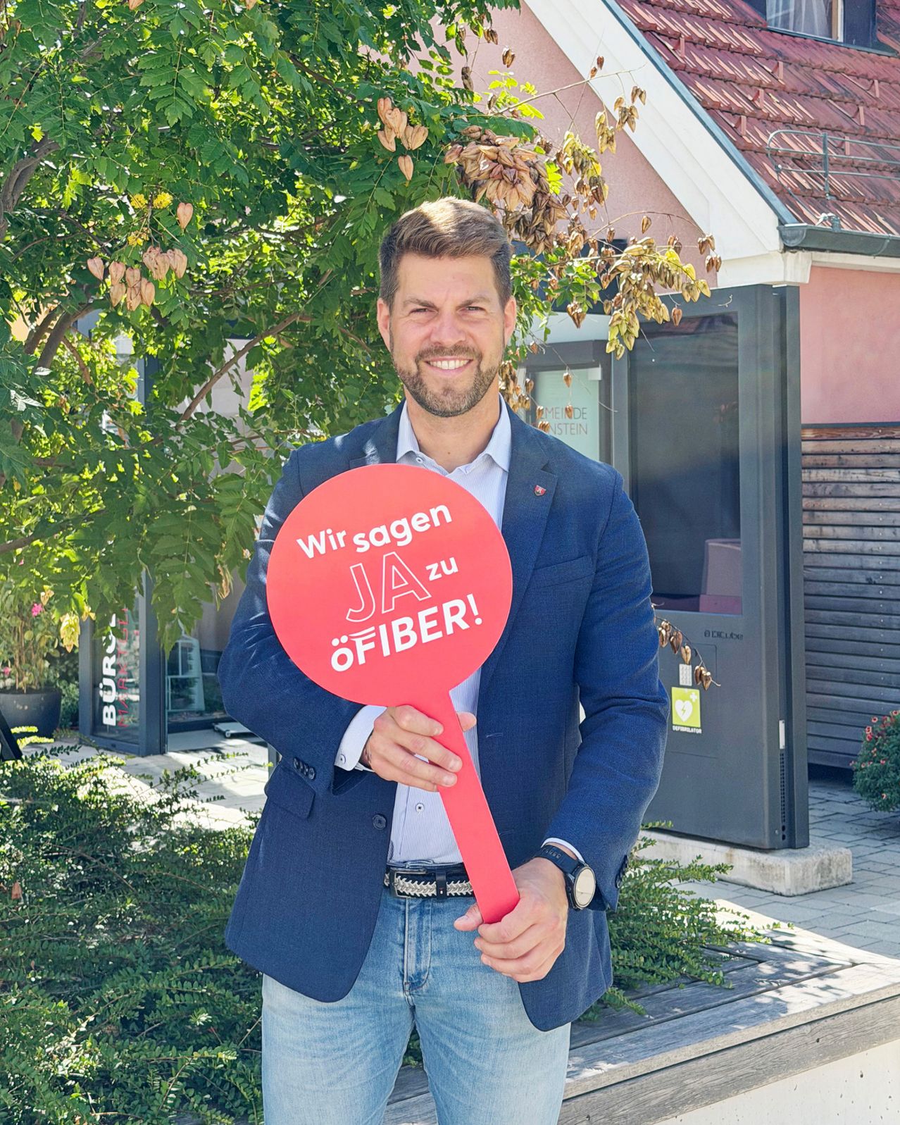 Ein Mann in einem blauen Anzug hält ein rotes Schild mit den Worten 'Wir sagen JA zu öFIBER!' vor einem Gebäude.