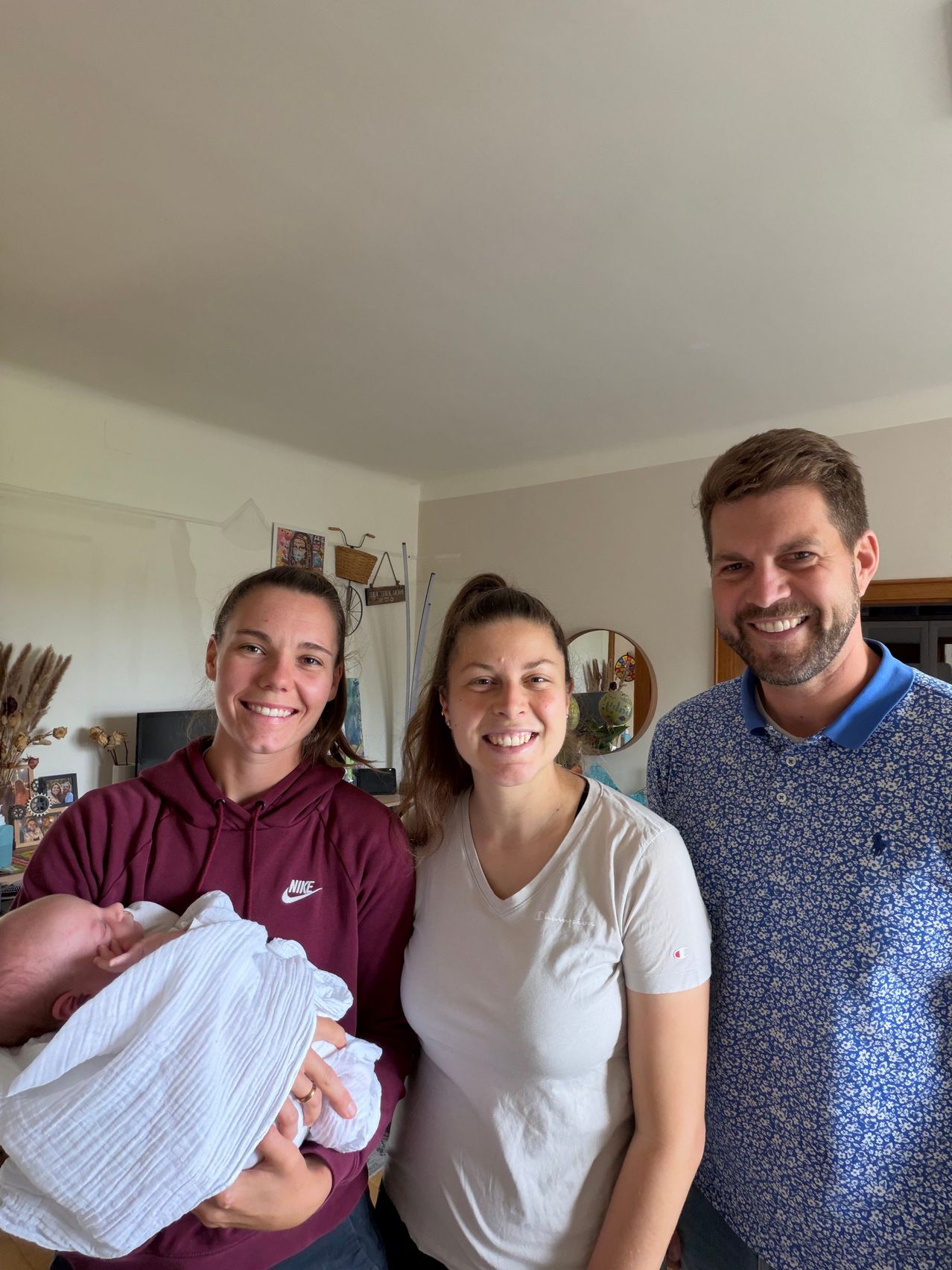 Drei Menschen lächeln und posieren für ein Foto in einem Zimmer. Eine Frau hält ein Baby.