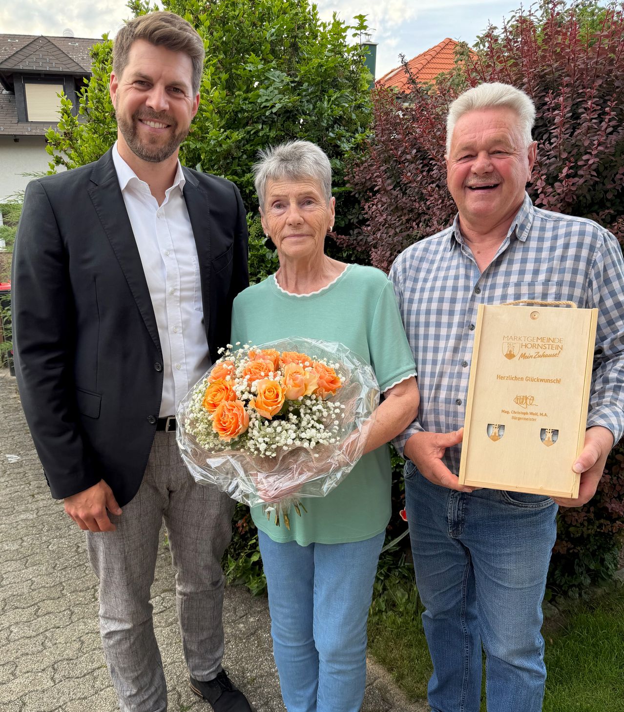 Drei Personen stehen lächelnd draußen, einer hält Blumen und ein Zertifikat. Die Frau hält einen Blumenstrauß, während zwei Männer ein Zertifikat halten. Hinter ihnen sind Bäume und ein Haus zu sehen.