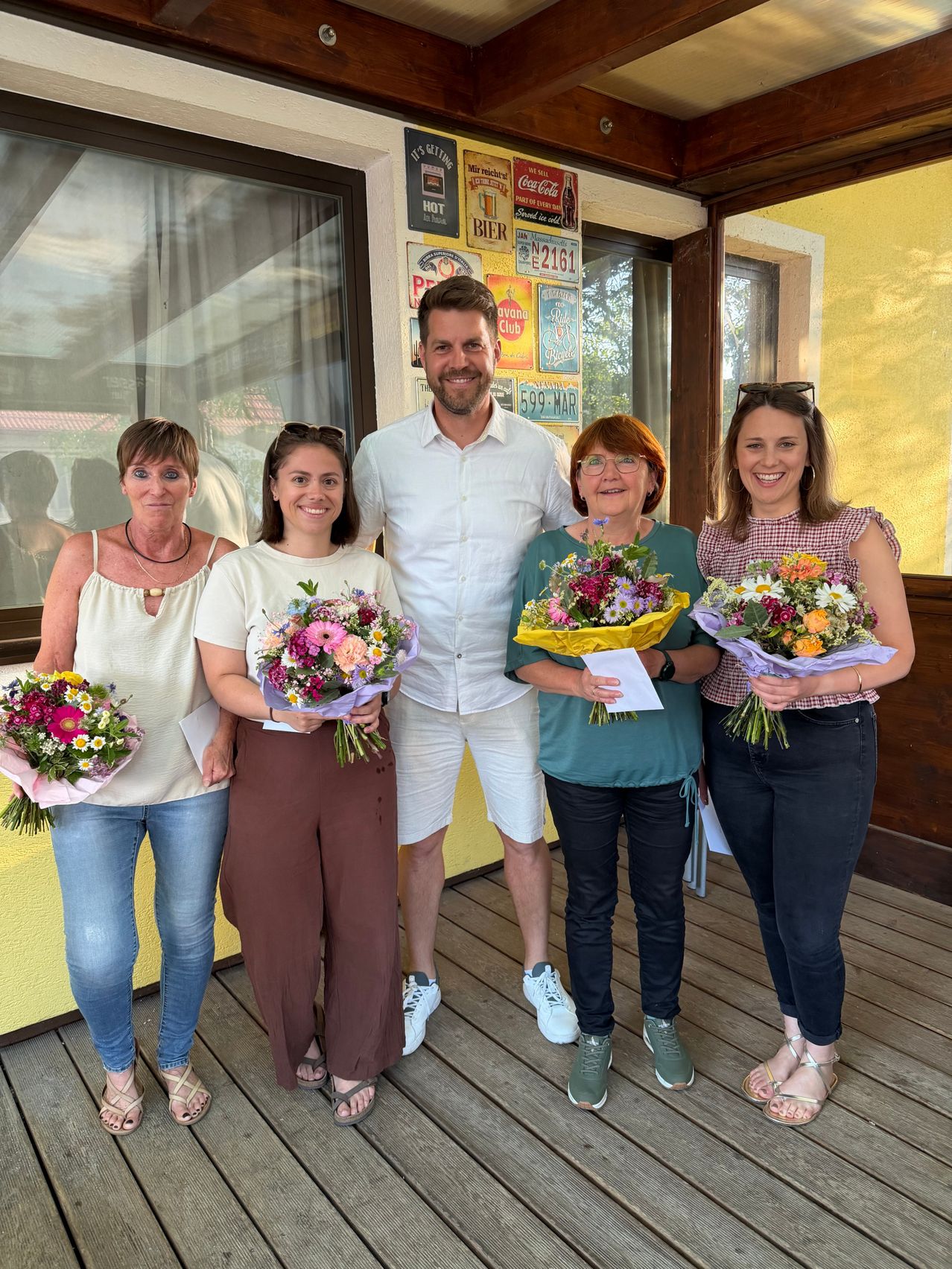 Fünf Personen stehen lächelnd auf einer Veranda mit gelber Wand. Ein Mann in einem weißen Hemd steht zwischen zwei Frauen, die Blumen halten. Eine weitere Frau mit einem Armband hält einen Blumenstrauß und Papier. Ein Coca-Cola-Schild ist an der Wand angebracht.