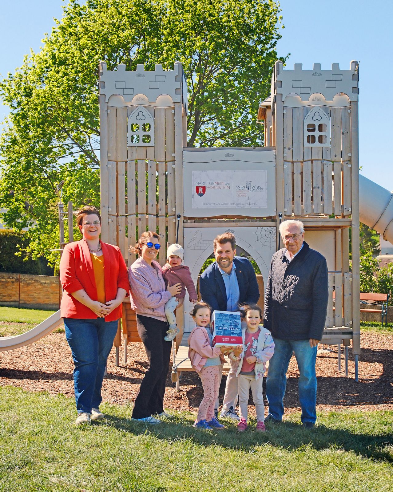 Eine sechsköpfige Familie, darunter zwei Kinder, steht vor einem Spielplatz mit einer großen Burgstruktur und einer Rutsche. Ein Mann hält eine Box und lächelt.