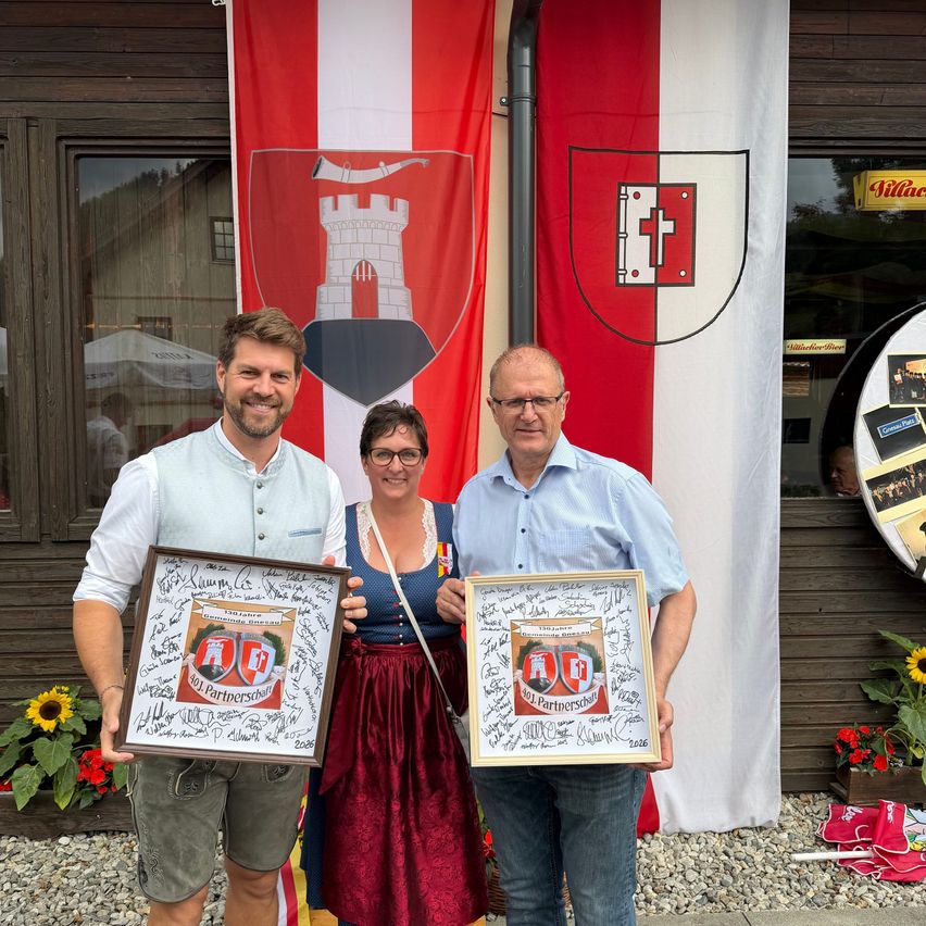 Three people hold framed certificates in front of a German flag. A man on the left holds a certificate. A woman in the middle wears a traditional dress and a man on the right holds a certificate. A building is behind them with flowers in front.