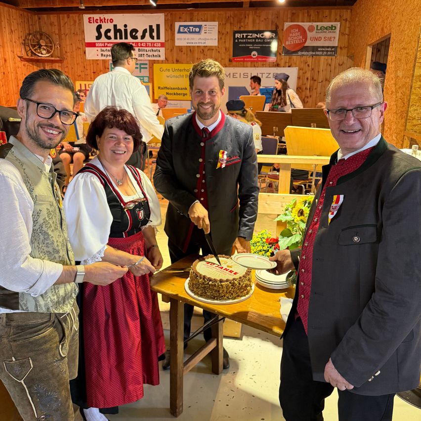Four people are standing around a table, smiling. Two men and a woman in traditional clothing are cutting a cake. There are plants and a banner in the background.