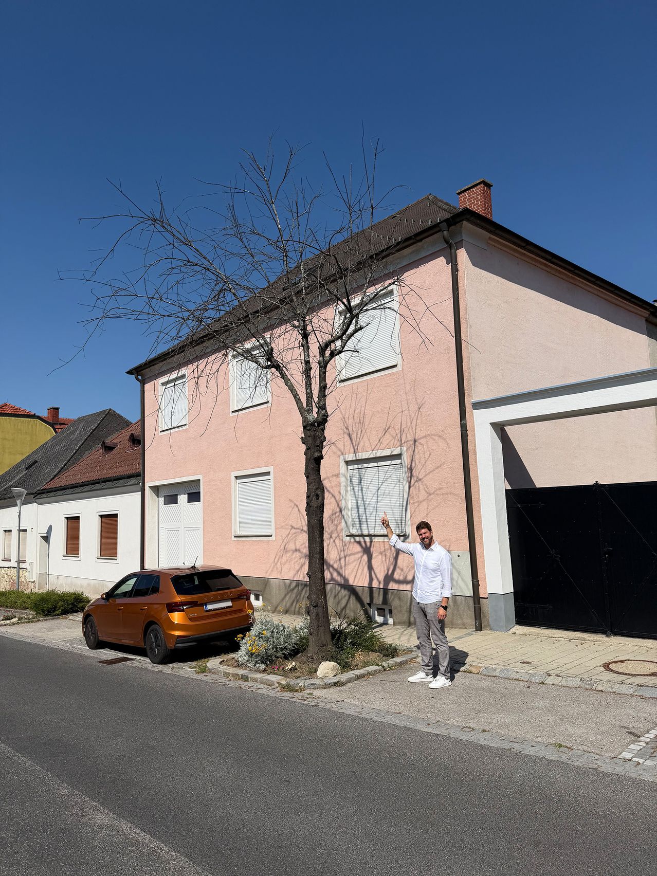 A man is standing next to a pink house with white shutters and a car parked in front.