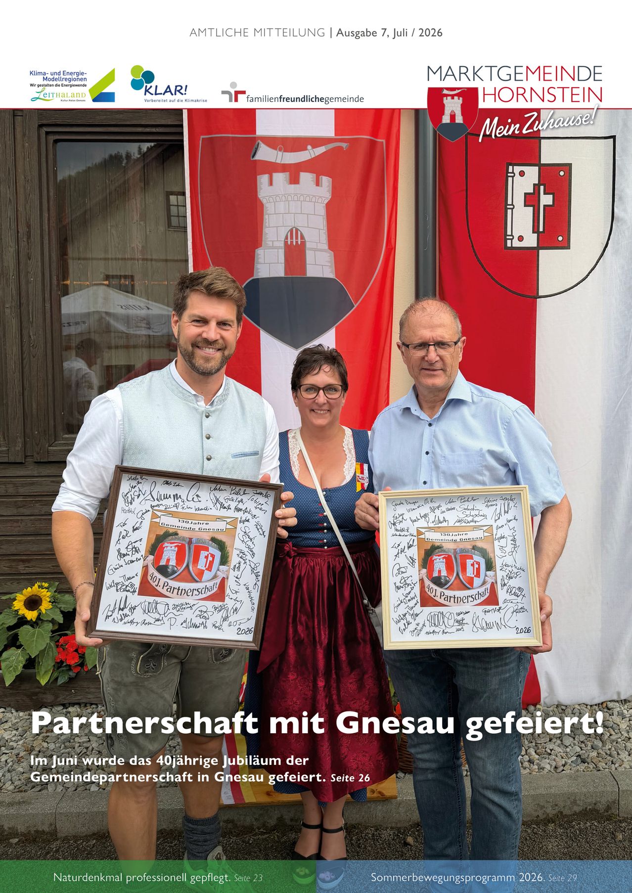 Three individuals pose with framed certificates in front of a German town flag. The man on the left wears a white shirt and suspenders. The woman in the middle dons a traditional dress. The man on the right wears glasses and a light blue shirt. The text reads 'Partnerschaft mit Gnesau gefeiert!'.