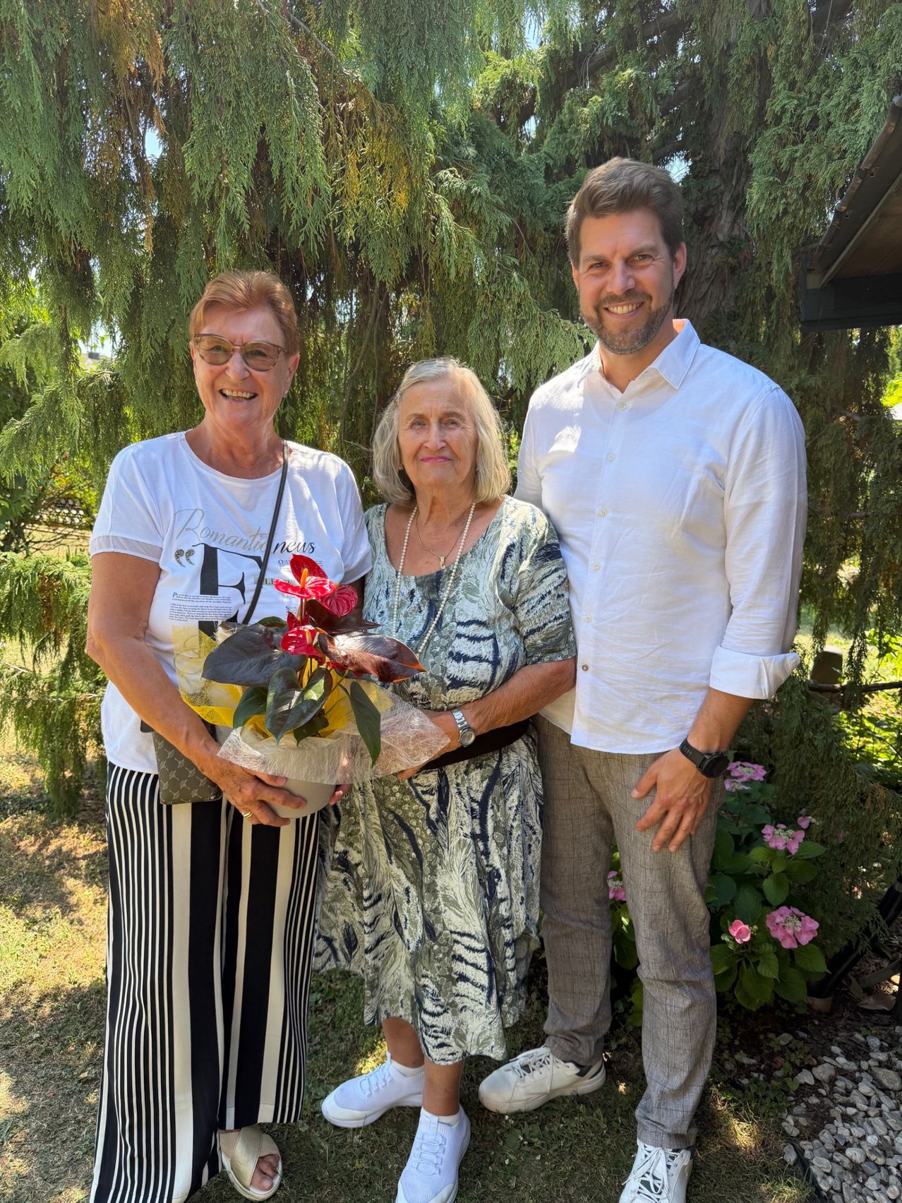 Three people stand in a garden, smiling. A woman holds a bouquet of red flowers, while a man and an elderly woman stand beside her. Trees and flowers are behind them.