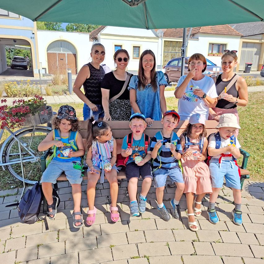 A group of people, including adults and children, are sitting on a bench under a green umbrella. They are eating ice cream and posing for a photo. Behind them are buildings, a bicycle, and a trash bin.