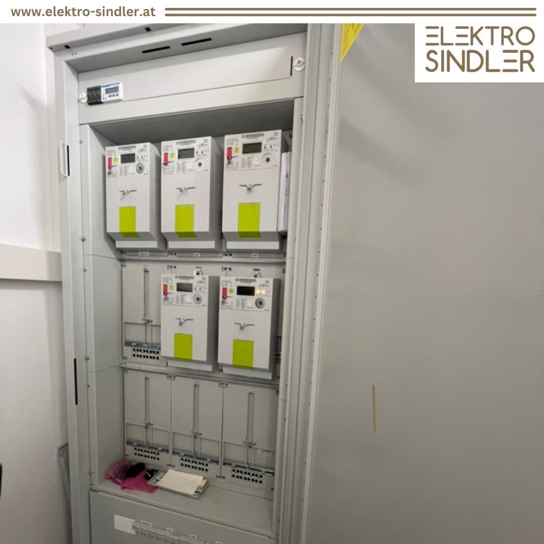 Ein offenes Schaltschrank zeigt mehrere elektronische Zähler mit gelben Etiketten, an einer weißen Wand. Der Text 'ELEKTRO SINDLER' befindet sich in der oberen rechten Ecke.