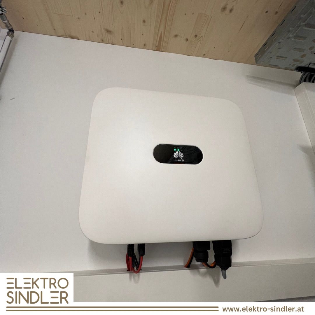 Ein weißer Huawei-Wechselrichter mit einem schwarzen Panel ist an einer weißen Wand mit einer Holzdecke montiert. Der Wechselrichter hat rote und schwarze Kabel angeschlossen. Das Bild hat einen Wasserzeichen mit dem Text 'ELEKTRO SINDLER' und einem Website-Link.