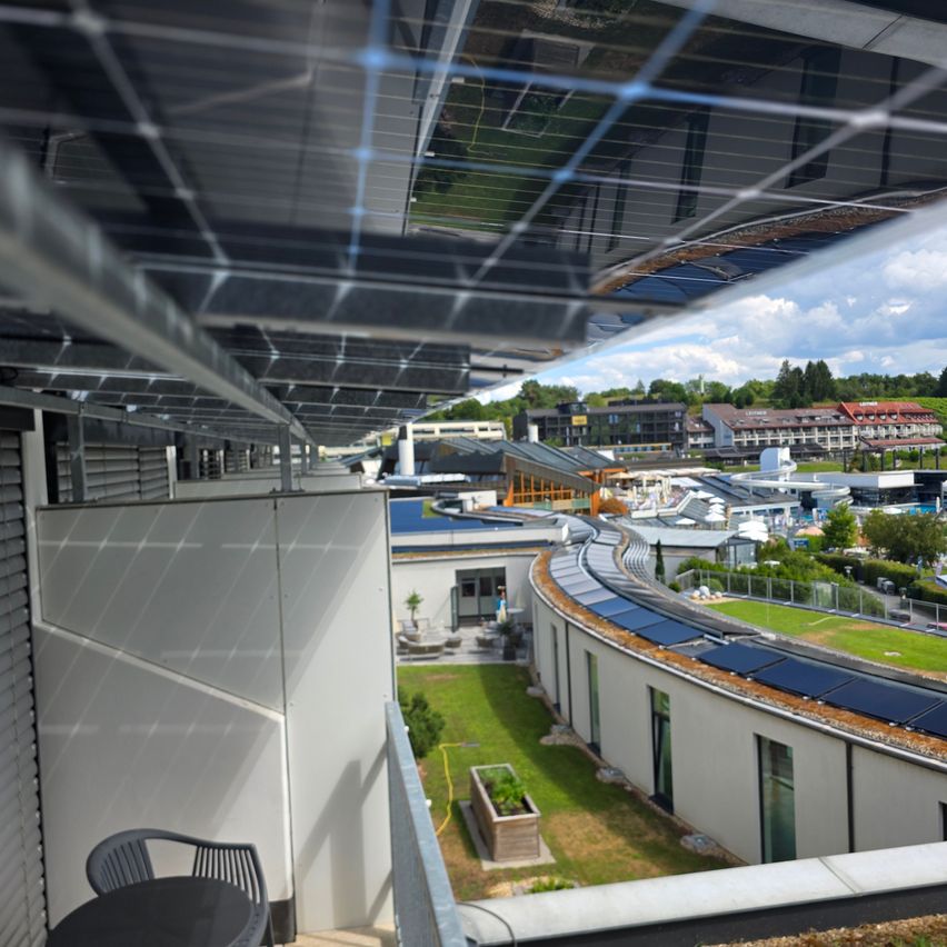 Ein Balkonblick auf ein modernes Gebäude mit Solarmodulen auf dem Dach, umgeben von Grünflächen und einem klaren Himmel.