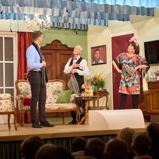 Drei Personen stehen auf der Bühne und spielen eine Szene. Einer ist in einem Anzug, einer in einem farbenfrohen Kleid und einer in einer Weste. Hinter ihnen steht eine Couch mit einem Kissen, ein kleiner Tisch mit Blumen und ein gerahmtes Bild an der Wand.