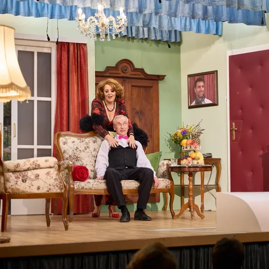 Eine Frau sitzt auf dem Schoß eines Mannes auf der Bühne. Ein Bild eines Mannes ist an der Wand und eine Lampe ist links. Ein Blumenvase steht auf dem Tisch.