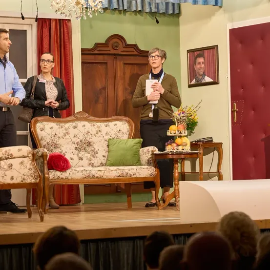 Drei Personen stehen auf einer Bühne vor einer roten Tür. Eine hält ein Papier, eine andere steht neben einer Couch mit einer roten Blume und eine dritte steht bei einem Tisch mit einem Blumenarrangement. Das Publikum beobachtet von vorne.