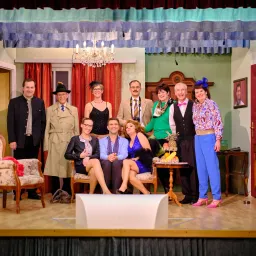 Eine Gruppe von Erwachsenen, wahrscheinlich Schauspieler, gekleidet in Vintage-Kleidung, posiert für ein Foto auf einer Bühne. Die Bühne ist mit einem Tisch, einer Laterne und einem Bilderrahmen dekoriert.