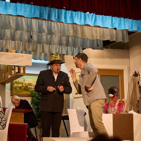 Ein Mann in Hut und Anzug steht und raucht eine Zigarette, während ein anderer Mann mit Sonnenbrille neben ihm steht. Im Hintergrund sitzt eine Frau am Tisch mit einer Blume und trinkt.