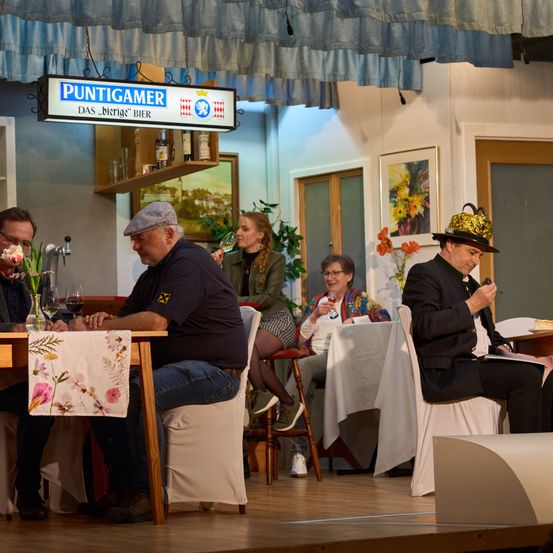 Fünf Erwachsene sitzen in einer Bühnenszene um einen Tisch. Einer trinkt aus einem Glas. Ein Schild steht 'Puntigamer' und 'Das Burgische Bier'.