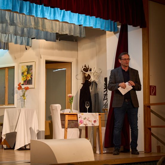 Ein Mann steht auf der Bühne, hält ein Papier in der Hand und trägt eine Brille. Hinter ihm steht ein kleiner Tisch mit einem Blumentopf und einem Weinglas. Links befindet sich ein Tisch mit einer weißen Tischdecke und Blumen. Ein Bilderrahmen und eine Tür sind an der Wand.