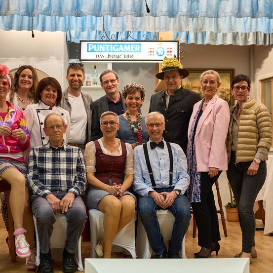 Eine Gruppe von Menschen posiert für ein Foto in einem Raum mit Holzboden. Über ihnen steht ein Schild mit der Aufschrift Puntigamer.