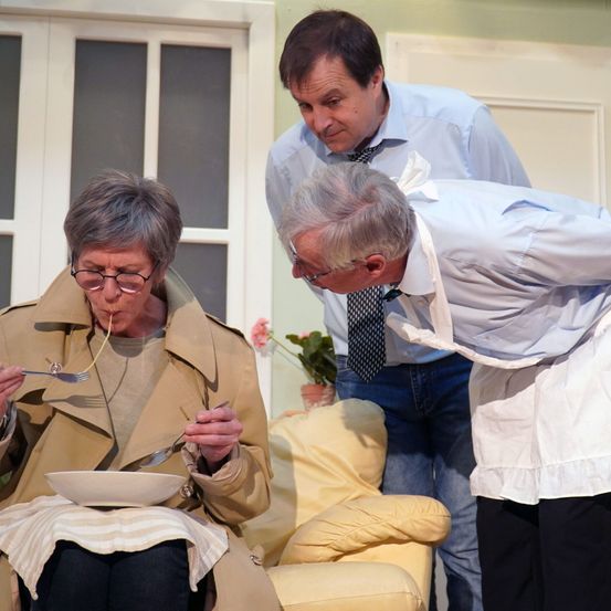 Drei Erwachsene in einem Raum. Eine Frau mit Brille sitzt auf einem Sofa und isst Nudeln. Ein Mann beugt sich über sie, während ein anderer hinter ihm steht.