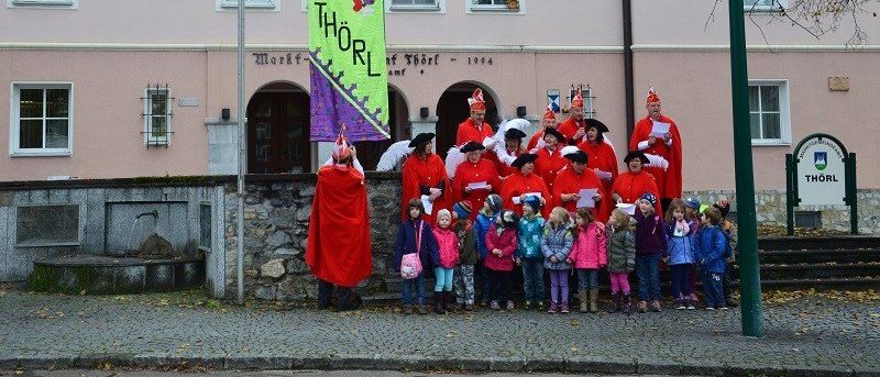 Eine Gruppe von Menschen in roten Kostümen und Hüten steht vor einem rosa Gebäude mit einem Banner, auf dem 'F.K. THORL' steht. Vor ihnen stehen Kinder.