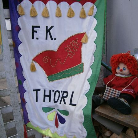Ein buntes Banner mit den Buchstaben F.K. Thorl und einem roten Hutdesign. Eine rothaarige Puppe sitzt daneben.