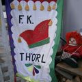 Ein buntes Banner mit den Buchstaben F.K. Thorl und einem roten Hutdesign. Eine rothaarige Puppe sitzt daneben.