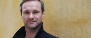 Ein Mann mit Bart und Schnurrbart steht vor einer gelben Wand und lächelt in die Kamera. Er trägt einen dunkelblauen Mantel.