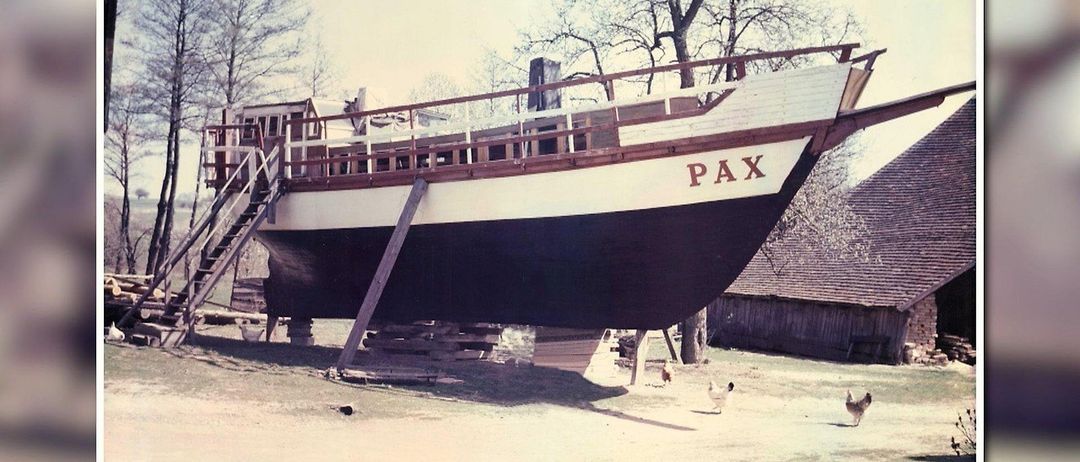 Ein großes Boot namens Pax wird auf einer Plattform in einem Hof gehoben, mit einem Scheunen und Bäumen im Hintergrund. Hühner gehen auf dem Boden.