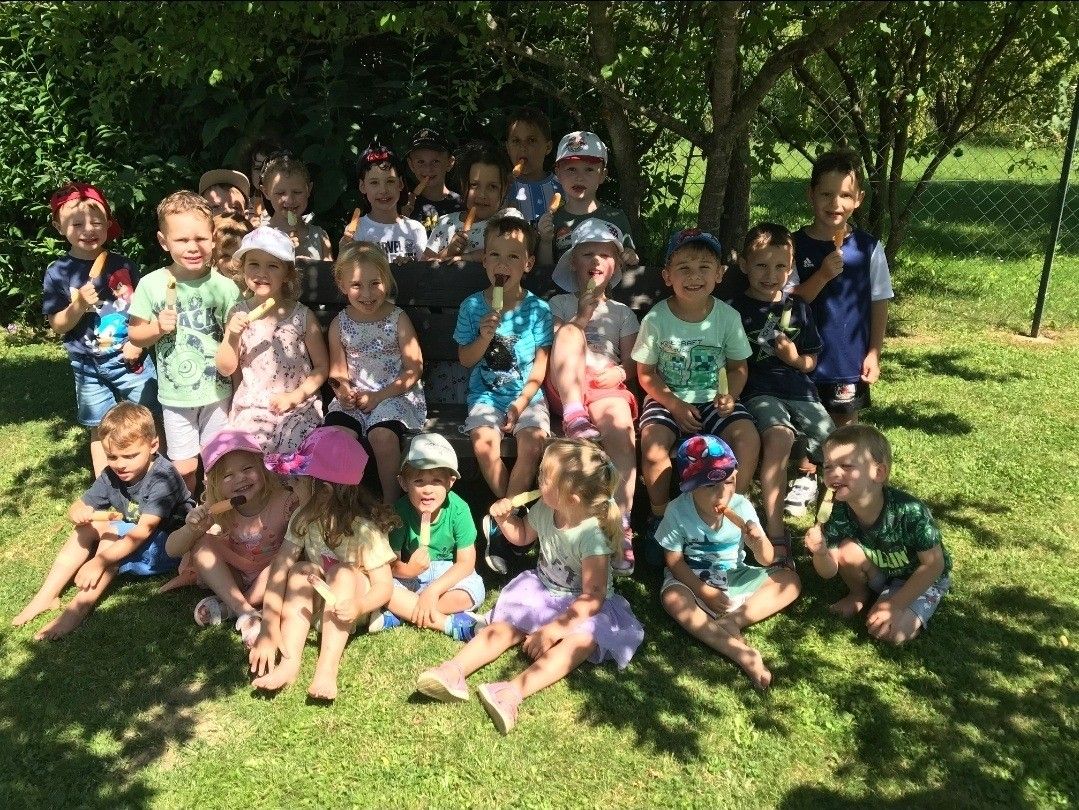 Eine Gruppe von Kindern, die auf Gras sitzen und Eislutscher halten. Einige tragen Hüte und Turnschuhe. Der Hintergrund hat Bäume und einen Zaun.