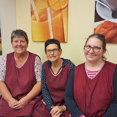 Drei Frauen in burgunderfarbenen Schürzen sitzen zusammen und lächeln. Hinter ihnen zeigt eine Wand einen Plakat, ein Glas Orangensaft und ein Croissant.
