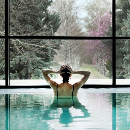 Eine Frau im Schwimmbad mit den Händen auf dem Kopf, die durch ein großes Glasfenster nach draußen schaut.