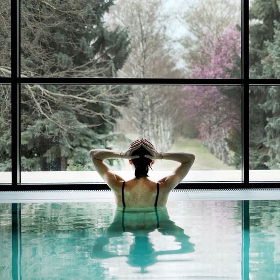 Eine Frau im Schwimmbad mit den Händen auf dem Kopf, die durch ein großes Glasfenster nach draußen schaut.