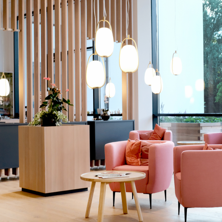 Ein moderner Loungebereich mit rosa Stühlen, einem Holztisch und hängenden Lampen. Der Raum hat Holzböden und ein großes Fenster mit Blick auf Bäume.