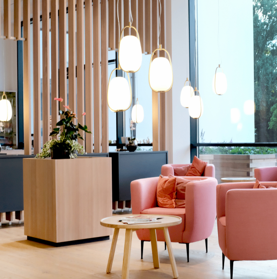 Ein moderner Loungebereich mit rosa Stühlen, einem Holztisch und hängenden Lampen. Der Raum hat Holzböden und ein großes Fenster mit Blick auf Bäume.