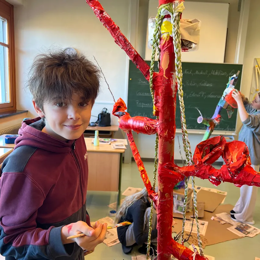 Ein Junge mit einer roten Kapuzenjacke malt in einem Klassenzimmer einen Baum mit einem Pinsel.