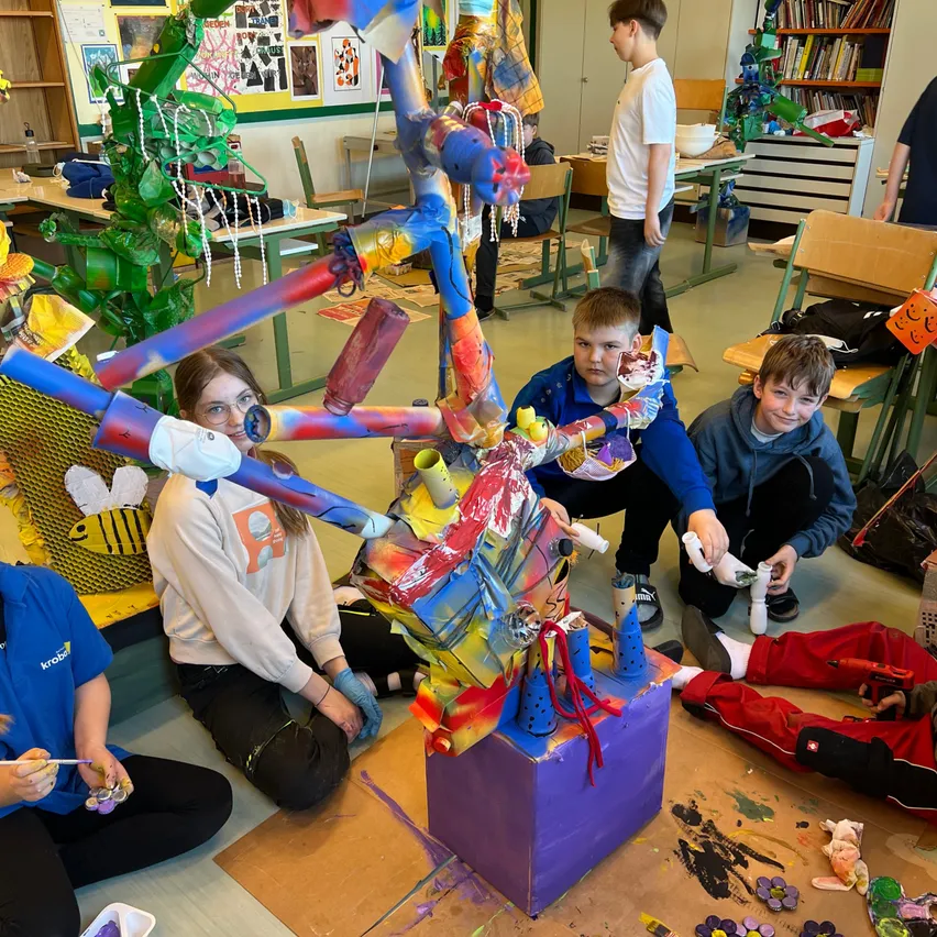 Eine Gruppe von Kindern malt eine bunte Skulptur in einem Klassenzimmer. Die Skulptur besteht aus verschiedenen Materialien wie Pappe, Plastik und Farbe.