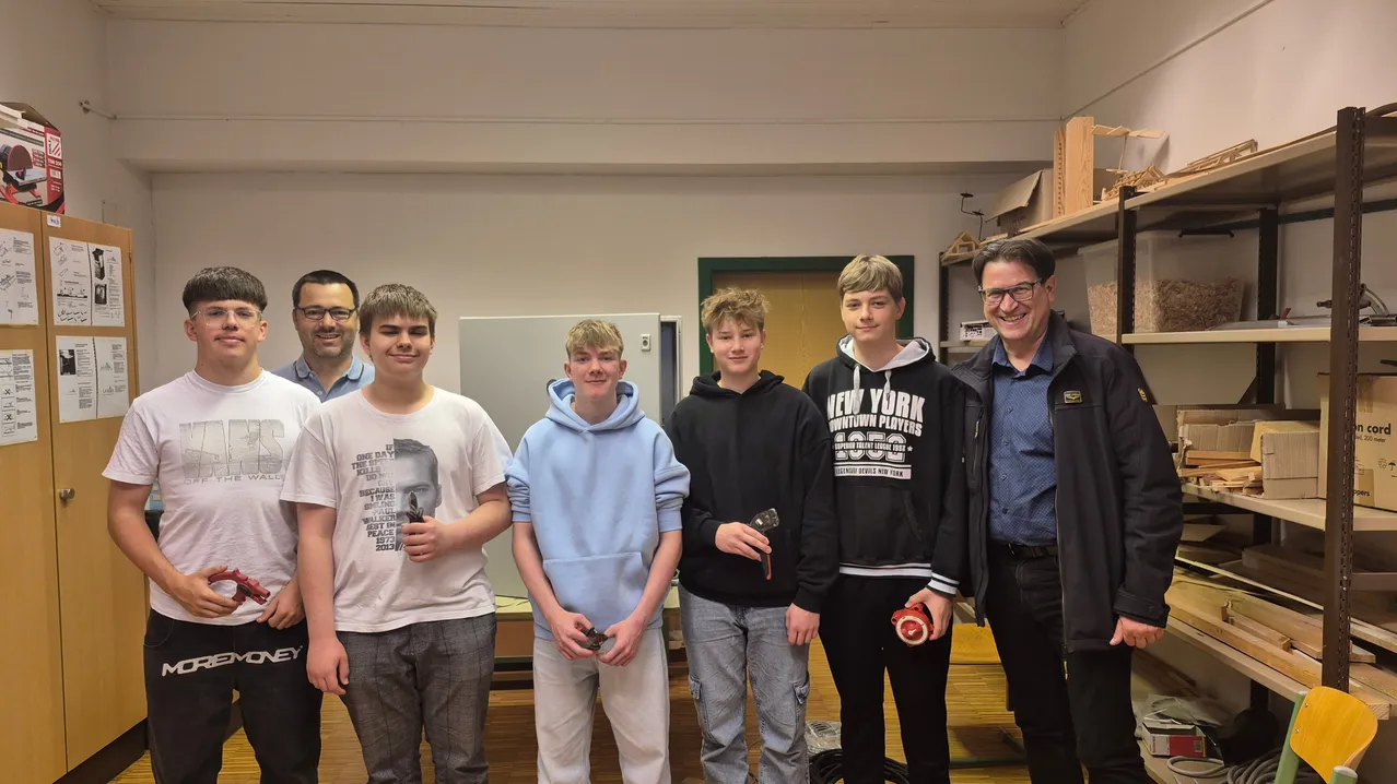 Fünf Jungen und ein Mann stehen lächelnd in einem Raum. Die Jungen halten Objekte in den Händen, und der Mann trägt eine Brille. Ein Tür, Regale und ein Holzboden sind im Hintergrund.