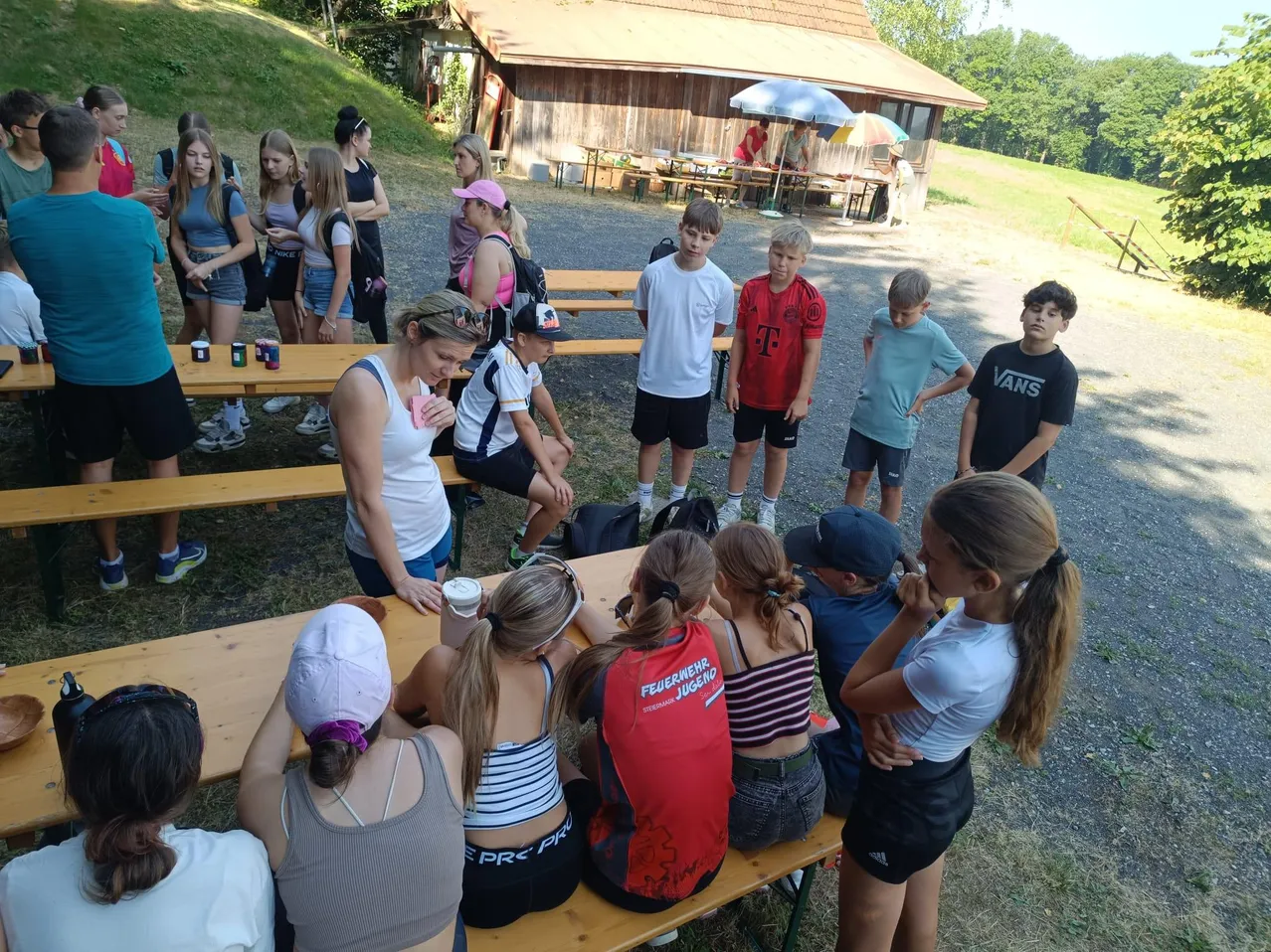 Eine Gruppe junger Menschen sitzt um einen Picknicktisch im Freien. Eine Frau steht und spricht die Gruppe an. Dahinter hat ein Gebäude Tische für eine Veranstaltung aufgestellt.