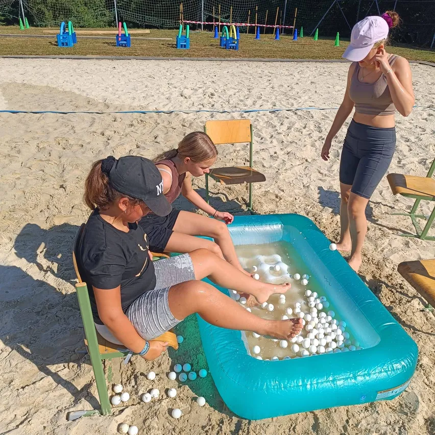Drei Frauen befinden sich am Strand, wobei eine in einem kleinen aufblasbaren Pool sitzt, der mit Ping-Pong-Bällen gefüllt ist. Eine weitere Frau sitzt neben dem Pool auf einem Stuhl, während eine dritte in der Nähe steht.