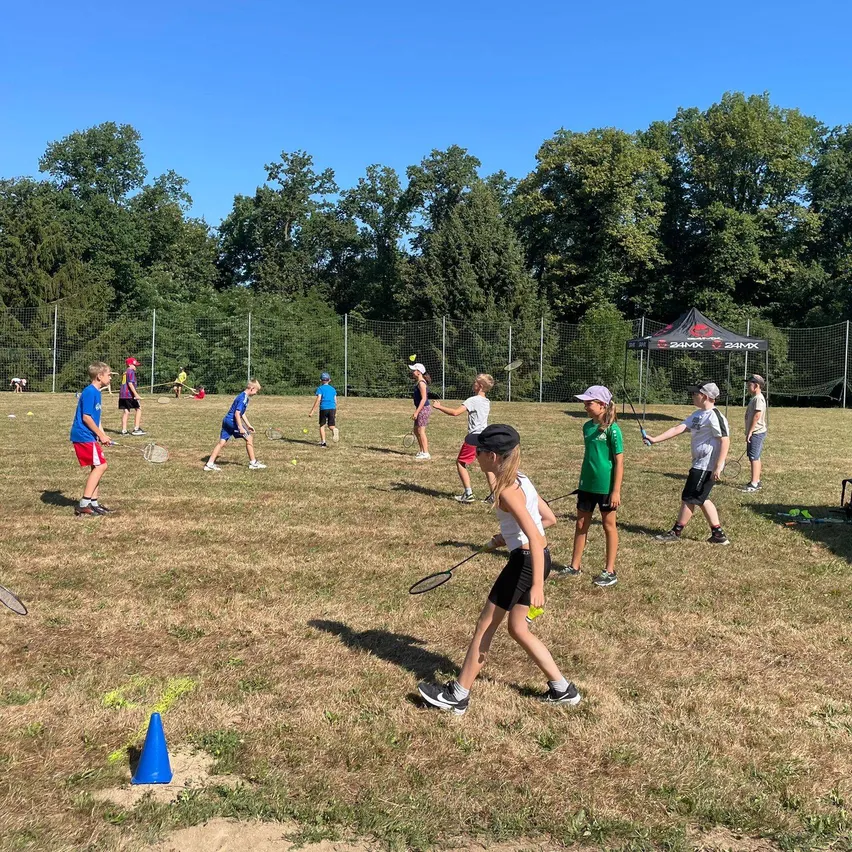 Eine Gruppe von Kindern spielt Badminton auf einem Grasfeld mit einem blauen Kegel, trägt Mützen und Turnschuhe. Bäume und ein Zelt sind im Hintergrund.