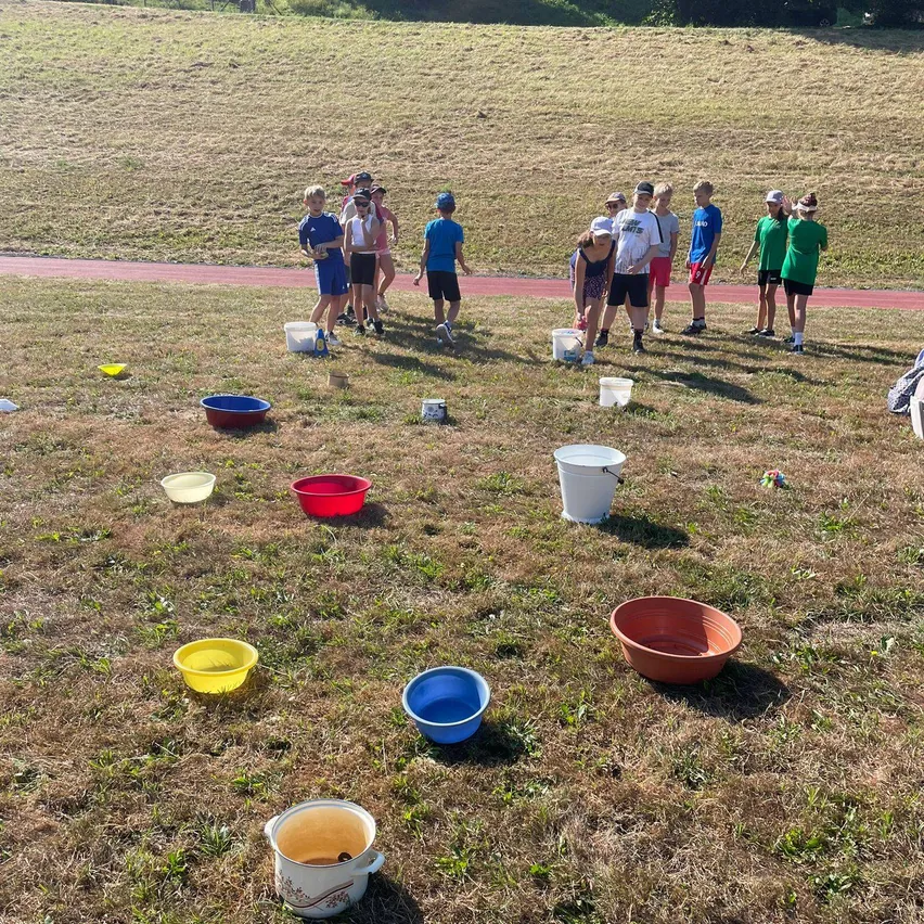 Eine Gruppe von Kindern steht auf einem Grasfeld, umgeben von Eimern und Schüsseln. Einige tragen Mützen und Turnschuhe. Eine Frau beugt sich vor, möglicherweise um etwas aufzuheben.
