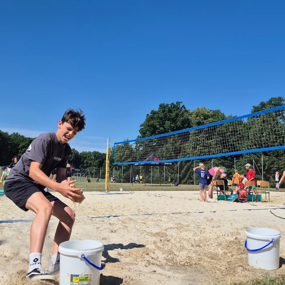 Ein junger Mann spielt Beachvolleyball auf einem sandigen Spielfeld, mit einem Netz davor und Menschen, die im Hintergrund stehen und sitzen.