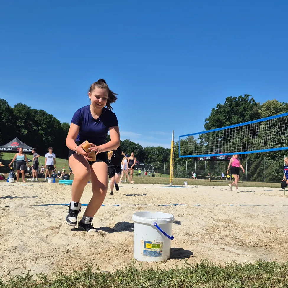 Eine Frau steht lächelnd im Sand in der Nähe eines Eimers. Im Hintergrund spielen Menschen Volleyball auf einem Sandplatz. Bäume und ein klarer Himmel vervollständigen die Outdoor-Atmosphäre.