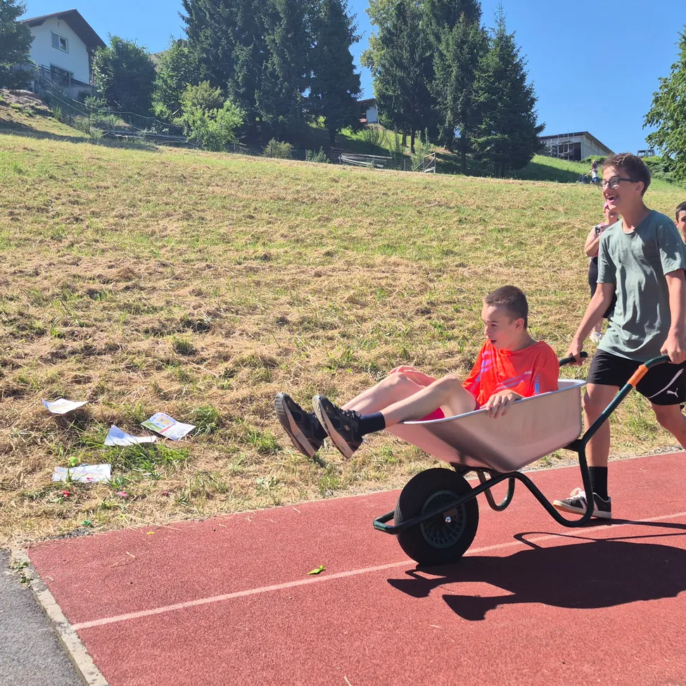 Ein junger Junge sitzt in einer Schubkarre, während ein anderer Junge ihn auf einer Laufbahn schiebt. Die Laufbahn ist von Gras umgeben. In der Ferne stehen Menschen.