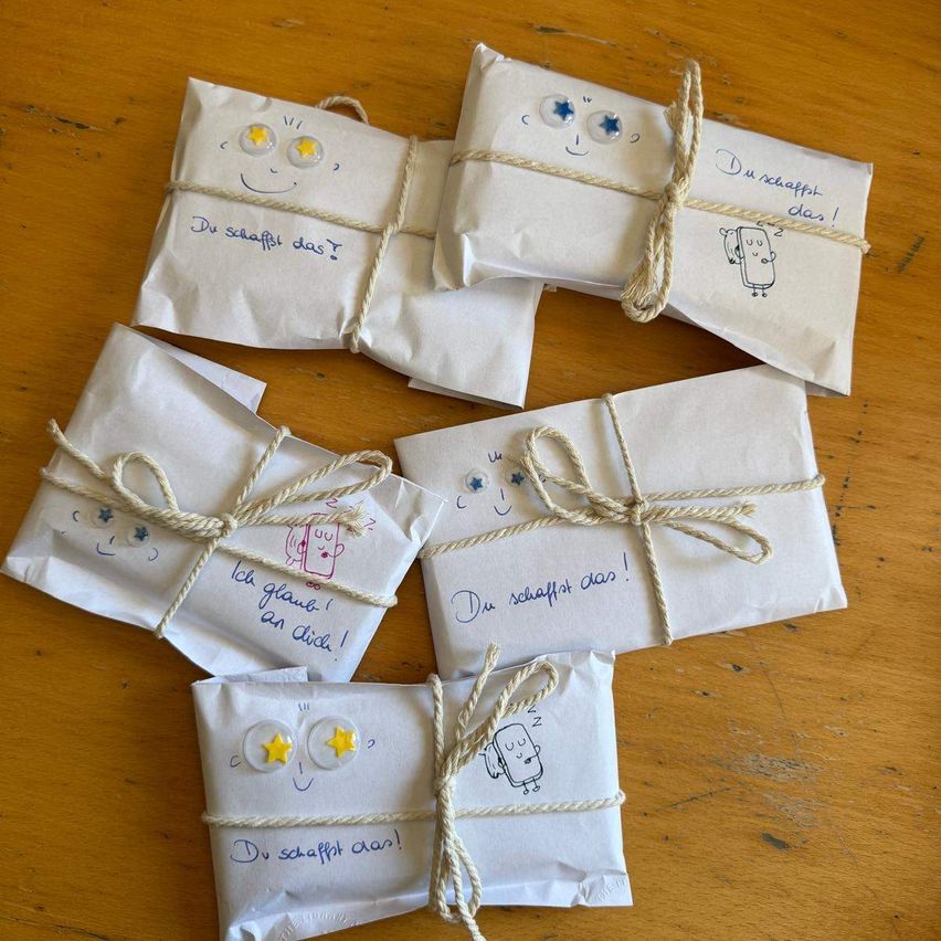 Mehrere kleine weiße Papierpakete, die mit Seil und Schnur gebunden sind, liegen auf einem Holztisch. Jedes Paket hat Zeichnungen und darauf geschriebene Phrasen.